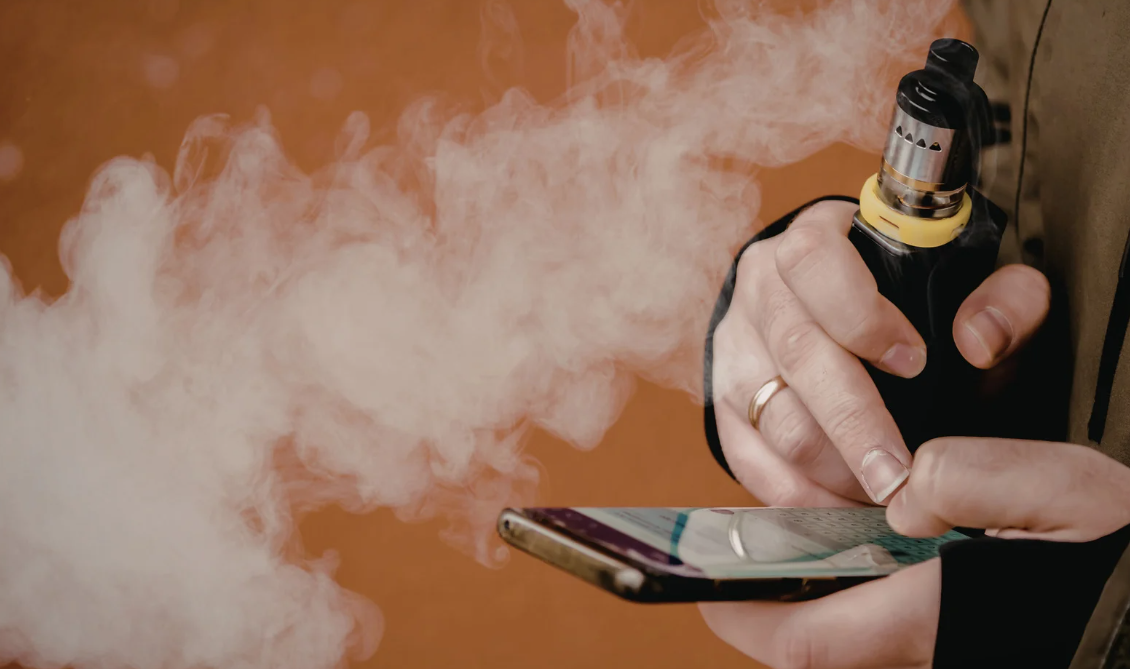 Pessoa segura um vape ao lado de um celular enquanto libera fumaça, em imagem sobre riscos do cigarro eletrônico.