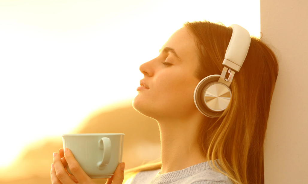 Mulher usando fones de ouvido enquanto segura uma xícara em ambiente iluminado, representando relaxamento com música.