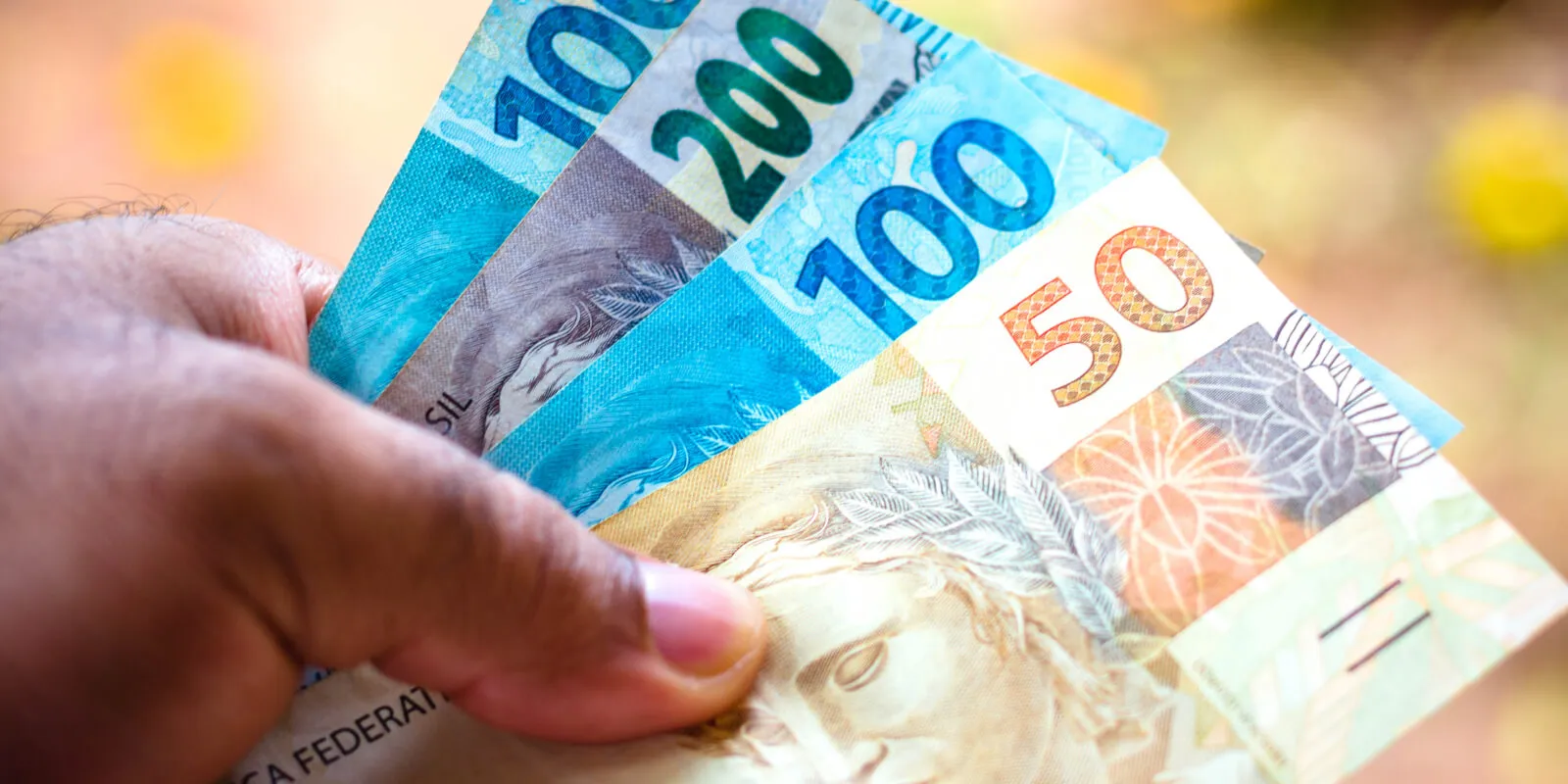 Mão segura cédulas de real em imagem sobre crédito, desconto em folha e empréstimo consignado.