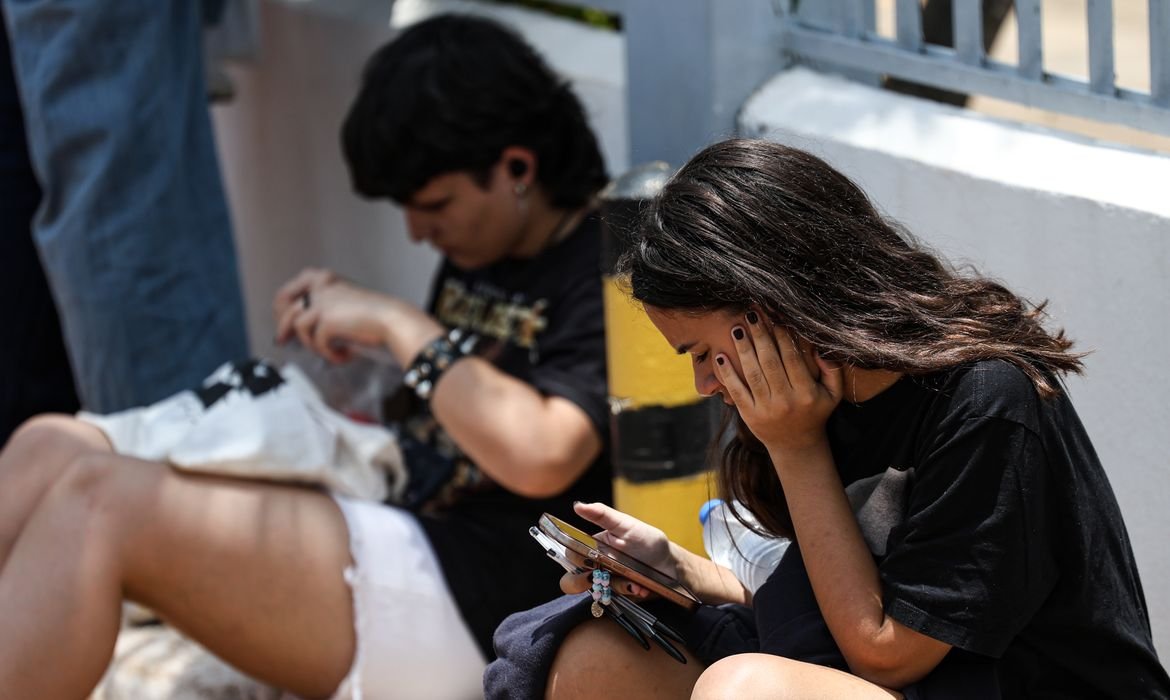 Estudantes aguardam sentados enquanto uma candidata acessa o celular em imagem usada sobre isenção do Enem 2026