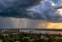 Inmet prevê chuva e trovoadas em Rondônia até Finados Composição ilustrativa de nuvens carregadas e chuva distante sobre cidade próxima a um rio, para notícia sobre chuva em Rondônia.